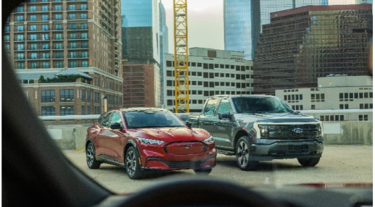 Urban landscape featuring a red Ford Mustang Mach-E and a silver electric Ford F-150 Lightning parked on a rooftop parking lot, with city skyscrapers and a construction crane in the background.