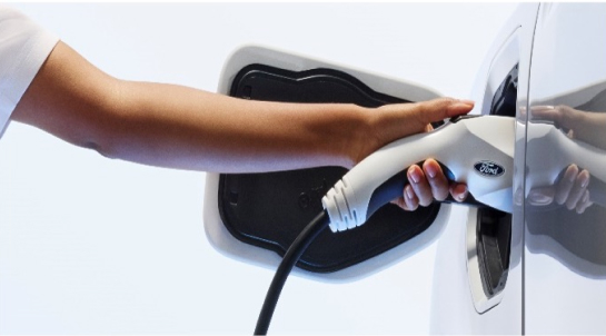 Close-up of a hand inserting a charging plug into the charging port of a white electric vehicle, featuring a Ford logo on the plug.