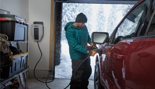 Man in winter clothing connecting a charging cable to a red electric vehicle in a snow-covered driveway, with a wall-mounted charging station visible in the background.
