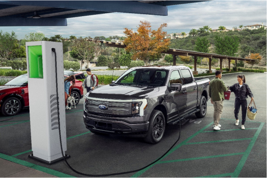 A gray electric pickup truck charging at a public charging station in a parking lot, with people walking nearby and green, tree-filled surroundings in the background.