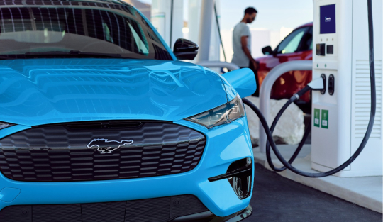 Man at a public charging station connecting a charging cable to a blue Ford Mustang Mach-E electric vehicle.