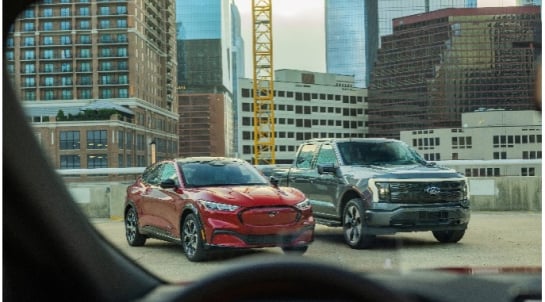 Urban landscape featuring a red Ford Mustang Mach-E and a silver electric Ford F-150 Lightning parked on a rooftop parking lot, with city skyscrapers and a construction crane in the background.
