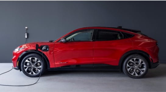 Red Ford Mustang Mach-E electric vehicle connected to a wall-mounted charging station, displayed against a gray background.