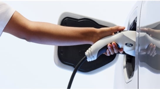 Close-up of a hand inserting a charging plug into the charging port of a white electric vehicle, featuring a Ford logo on the plug.
