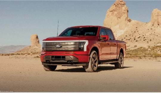 Red Ford F-150 Lightning electric pickup truck parked on a desert road with a rocky landscape in the background.