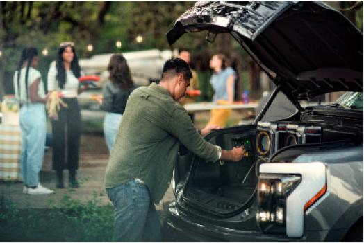 Man adjusting a speaker in the front trunk (frunk) of an electric vehicle, with a group of people chatting in the background at a park.