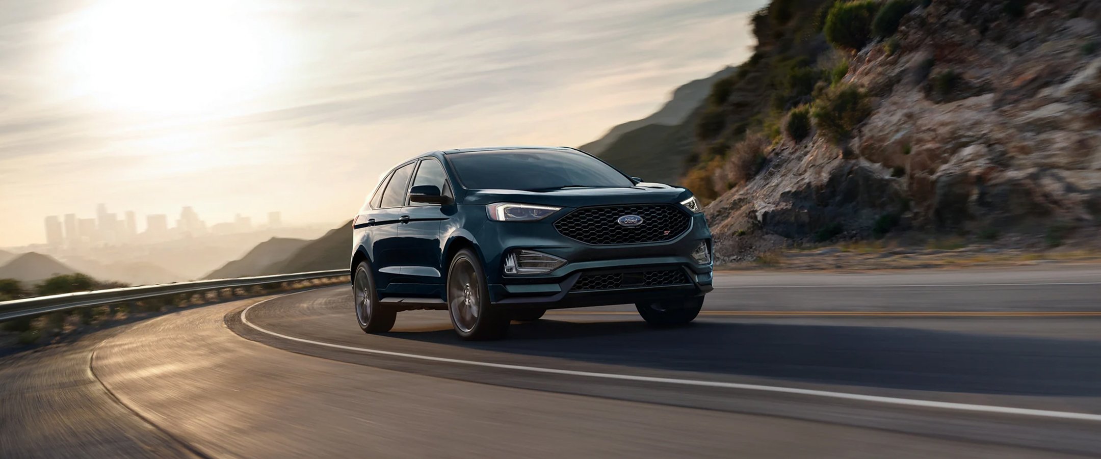 Blue 2023 Ford Edge driving on a windy mountainside road with a city landscape in the background.