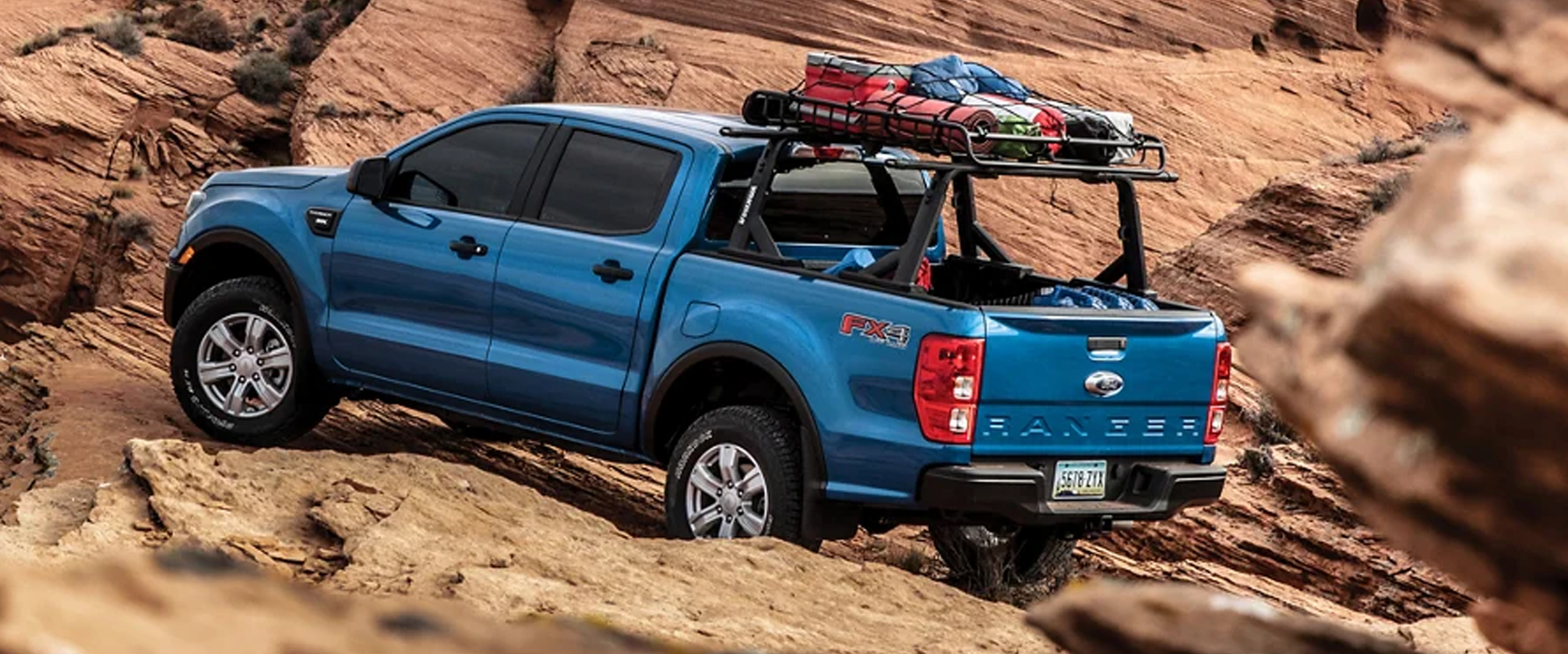 Blue 2023 Ford Ranger driving in the desert with a roof rack of camping supplies.