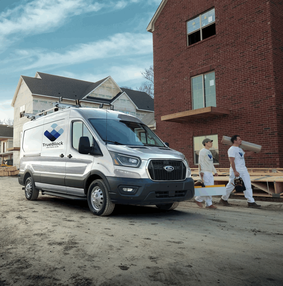 Ford Transit blanc avec échelle sur le toit, stationné près de travailleurs devant un bâtiment en briques.