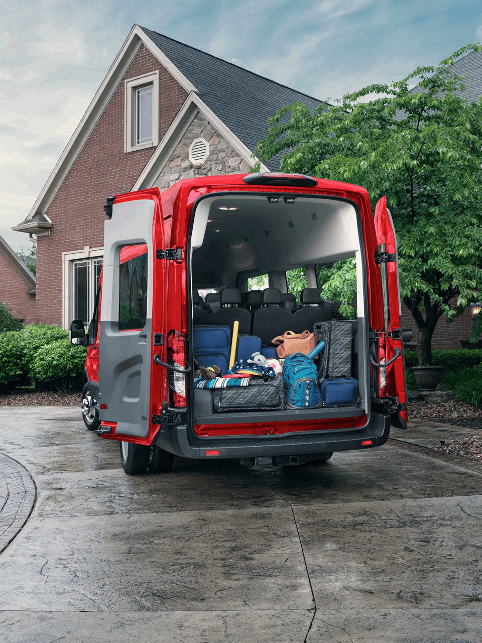 Ford Transit rouge aux portes arrière ouvertes, chargé de sacs et de matériel devant une maison.