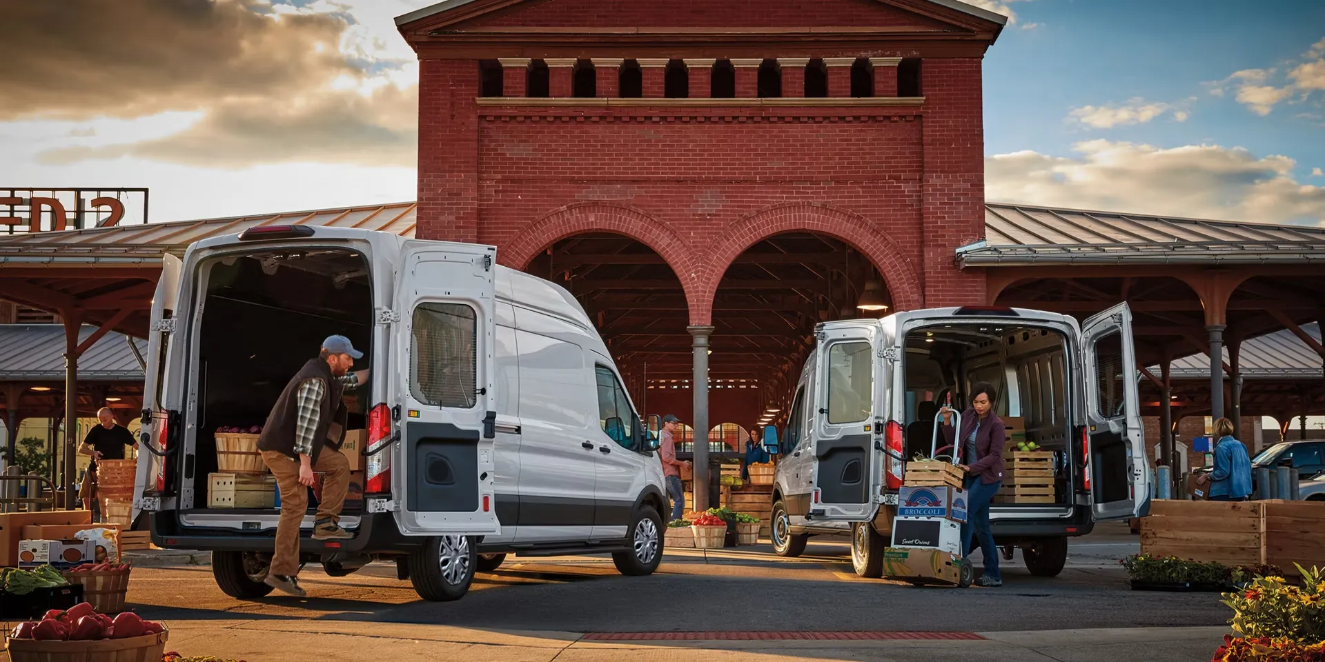 Deux fourgons Ford Transit blancs aux portes arrière ouvertes lors du déchargement de marchandises sur un marché.